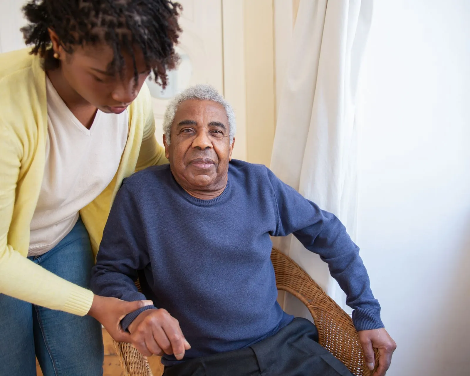 Caregiver helping an older adult stand from a chair in a sunlit living room