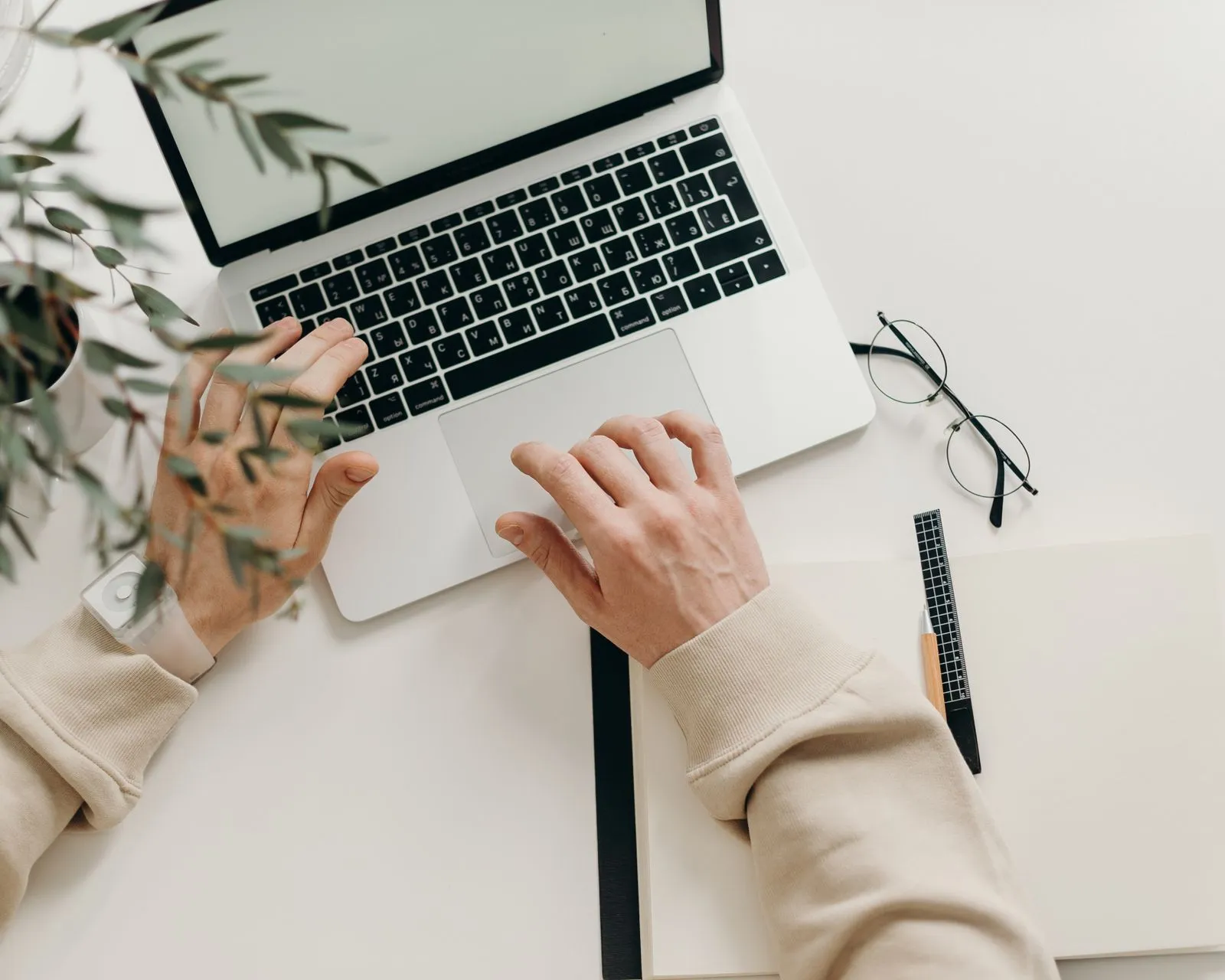 Hands using a laptop on a clean desk beside a notebook and glasses