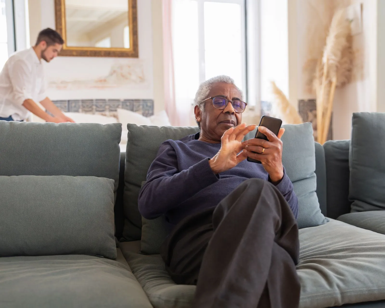 Caregiver making a bed in a bright home while an older adult sits nearby with a phone
